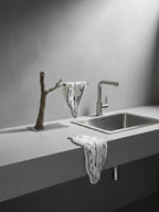 A DAMNART$ Dalí-style melting clock towel draped over a sculptural branch beside a modern stainless-steel sink under dramatic lighting.