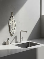 A DAMNART$ melting clock towel inspired by Dalí’s surrealist clocks, hanging on a minimalist grey kitchen wall beside a modern sink.