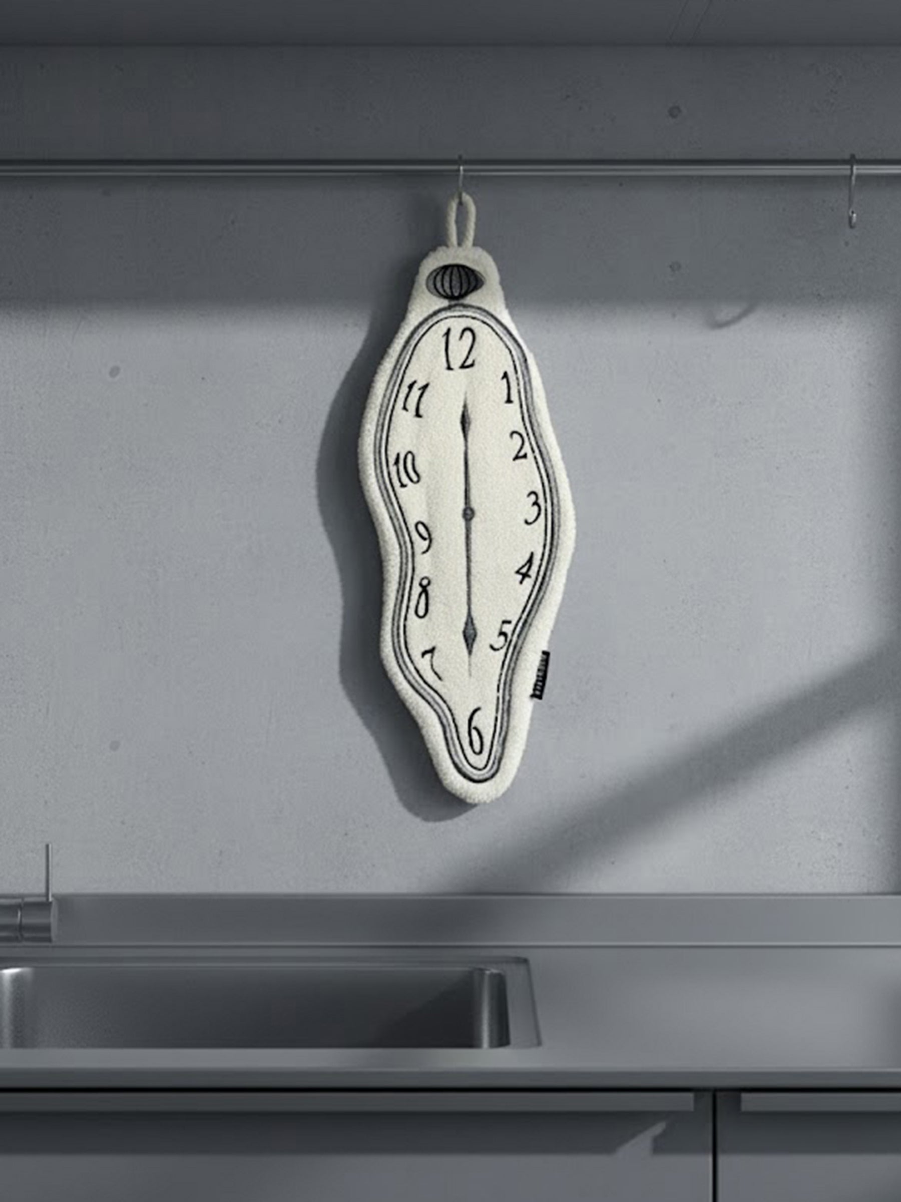 A DAMNART$ melting clock towel inspired by Dalí’s surrealist clocks, hanging on a minimalist grey kitchen wall beside a modern sink.