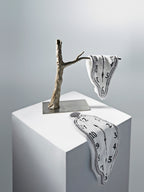 Two Dalí-inspired DAMNART$ melting clock towels displayed on a sculptural branch and white pedestal in a clean gallery-like setting.