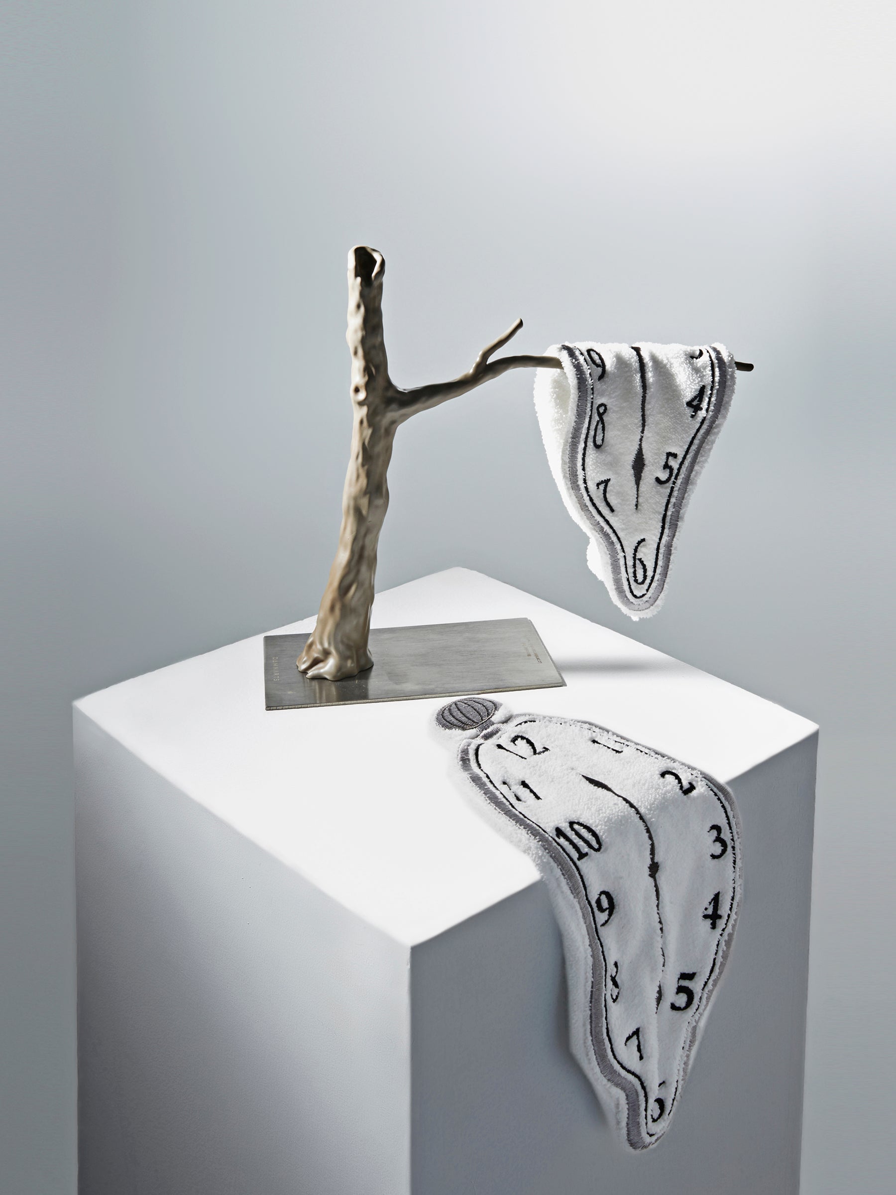 Two Dalí-inspired DAMNART$ melting clock towels displayed on a sculptural branch and white pedestal in a clean gallery-like setting.