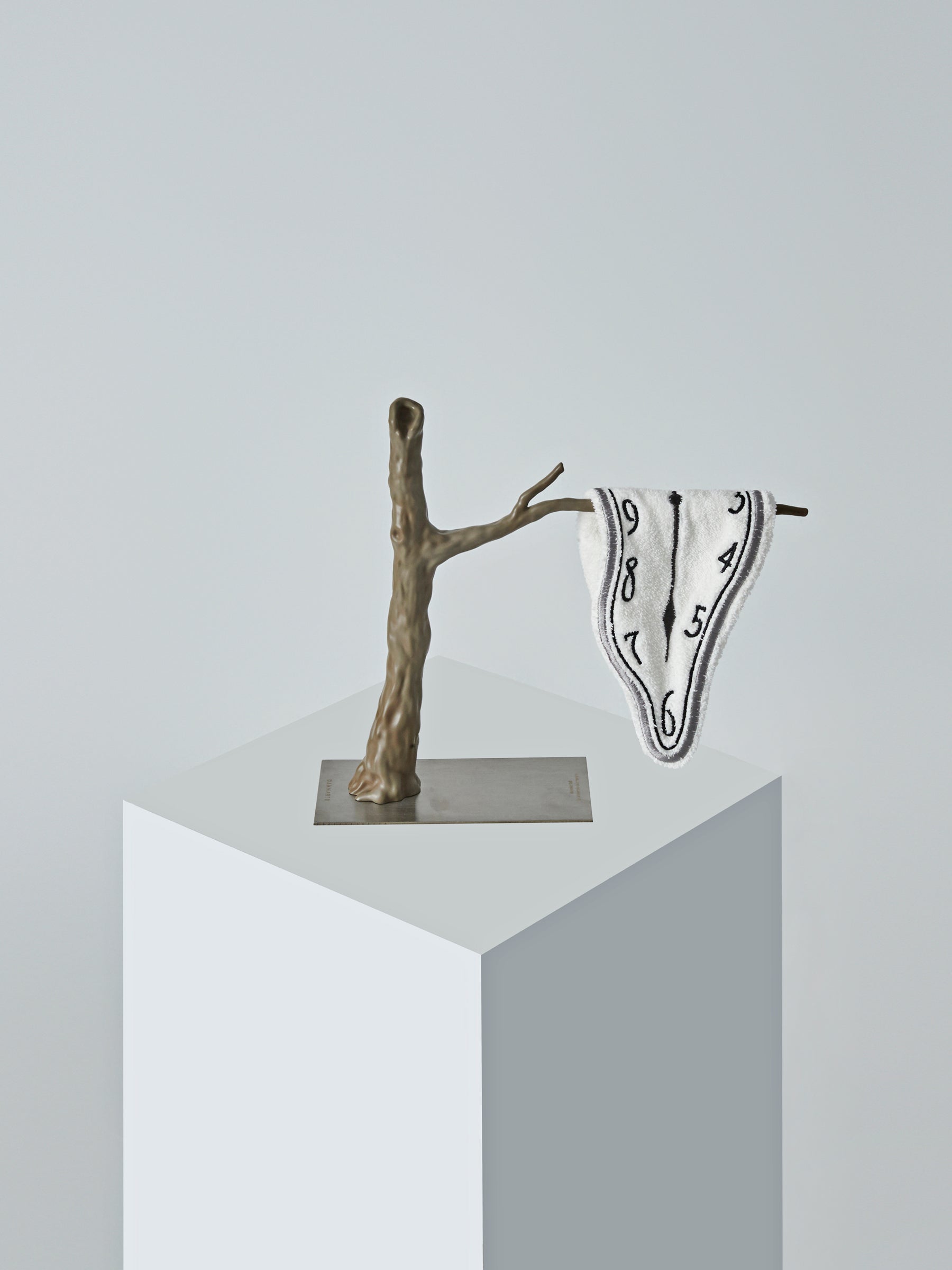 A single Dalí-style melting clock towel by DAMNART$ draped over a sculptural branch on a minimalist pedestal.