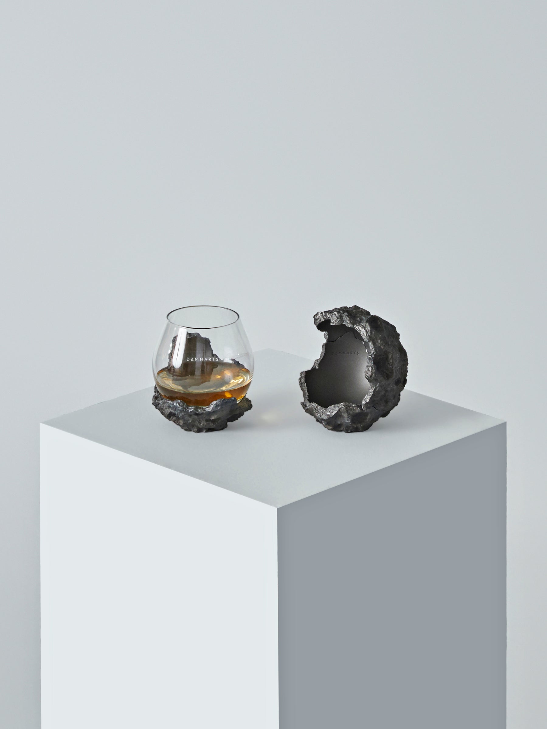 A DAMNARTS meteorite-style whisky glass set displayed on a minimalist white pedestal, featuring a textured meteorite base and a sculptural half-sphere holder.