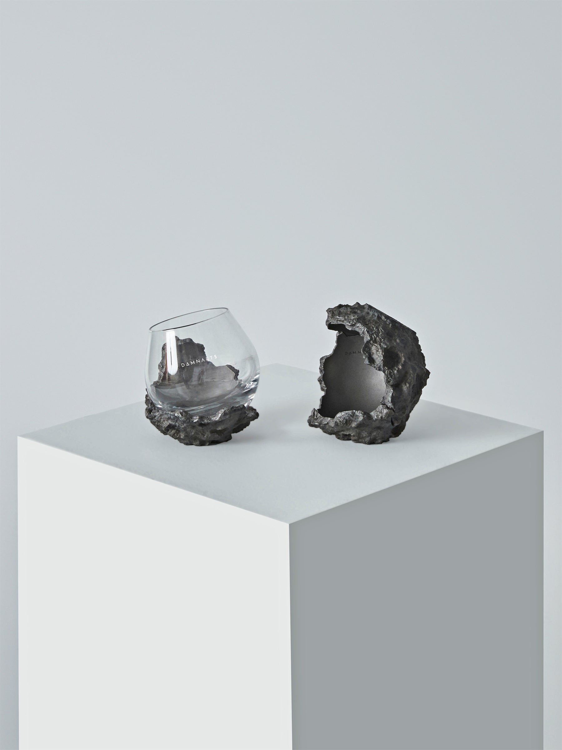 DAMNARTS meteorite whisky glass and cratered holder presented on a clean pedestal in a minimalist studio setting.